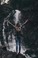 Rückansicht in voller Länge einer jungen Frau mit Rucksack, die die Hände hochhält, während sie in der Nähe des Wasserfalls steht foto