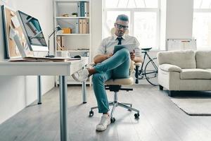 kabellose Technologie. Gut aussehender junger Mann in Hemd und Krawatte mit digitalem Tablet und lächelnd, während er im Büro sitzt foto