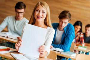 glücklich, der Beste zu sein. Fröhliche junge Frau, die weißes Papier hält und in die Kamera schaut, während sie an der Rezeption im Klassenzimmer sitzt foto