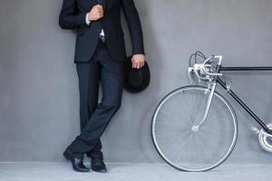 eleganter Stil. Nahaufnahme eines jungen Geschäftsmannes, der Hut hält und seine Jacke anpasst, während er neben seinem Fahrrad vor grauem Hintergrund steht foto