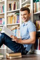 Studentin in der Bibliothek. Seitenansicht eines hübschen jungen Mannes, der ein Buch hält und in die Kamera lächelt, während er auf dem Boden sitzt und sich an das Bücherregal der Bibliothek lehnt