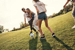 versucht zu gewinnen. Gruppe junger lächelnder Menschen in Freizeitkleidung, die einen schönen Sommertag genießen, während sie draußen Fußball spielen foto