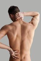 Schmerzen fühlen. Rückansicht eines jungen muskulösen Mannes, der seinen Rücken berührt, während er isoliert auf grauem Hintergrund steht foto