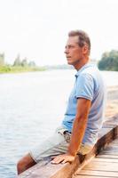 Tagträumen am Flussufer. Seitenansicht eines nachdenklichen reifen Mannes, der wegschaut, während er am Kai sitzt foto