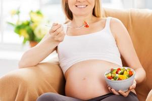 frischen Salat essen. Zugeschnittenes Bild einer glücklichen schwangeren Frau, die auf dem Stuhl sitzt und Salat isst foto