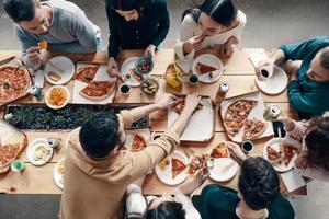 Abendessen unter Freunden. Draufsicht auf junge Leute in Freizeitkleidung, die Pizza pflücken, während sie drinnen eine Dinnerparty haben foto