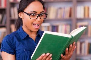 was für ein aufregendes buch begeisterte afrikanische studentin, die ein buch liest und den mund offen hält, während sie in der bibliothek steht foto