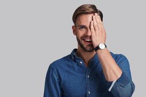 glücklich fühlen. hübscher junger Mann, der mit einem Lächeln in die Kamera blickt und das Auge mit der Hand bedeckt, während er vor grauem Hintergrund steht foto