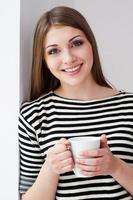 frischen Kaffee trinken. Schöne junge Frau in gestreifter Kleidung, die sich an die Wand lehnt und eine Tasse hält foto