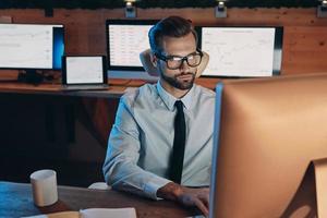 Selbstbewusster junger Mann, der am Computer arbeitet, während er lange im Büro bleibt foto