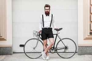 Bist du bereit für die Fahrt in voller Länge von einem hübschen jungen bärtigen Mann, der sich an sein Fahrrad lehnt und die Kamera anschaut, während er im Freien steht? foto