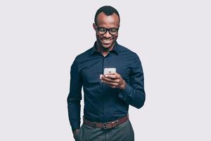 tolle Neuigkeiten von Freund. hübscher junger afrikanischer mann, der smartphone hält und es mit einem lächeln betrachtet, während er vor grauem hintergrund steht foto