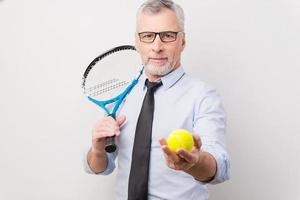 Machen Sie eine Pause Selbstbewusster älterer Mann mit grauem Haar in Hemd und Krawatte, der Tennisschläger hält und Tennisball ausstreckt, während er vor weißem Hintergrund steht foto