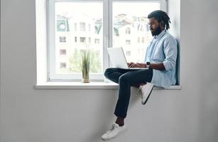 nachdenklicher junger afrikaner, der einen laptop benutzt, während er drinnen auf der fensterbank sitzt foto