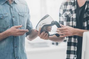 Probieren Sie die neue Welt der virtuellen Realität aus Nahaufnahme von zwei jungen Geschäftsleuten in Freizeitkleidung, die sich unterhalten, während einer von ihnen einem anderen ein VR-Headset gibt foto