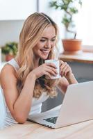 frischer Klatsch online. Schöne, fröhliche junge Frau, die lächelnd auf ihren Laptop schaut und eine Kaffeetasse hält, während sie zu Hause in der Küche sitzt foto