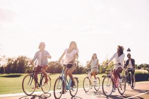 beste freunde und weiter gehts. Gruppe junger Leute, die Fahrräder entlang einer Straße fahren und glücklich aussehen foto