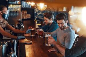 lächelnde junge Männer in Freizeitkleidung, die Bier trinken und sich zusammenschließen, während sie in der Kneipe sitzen foto