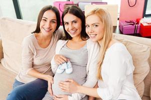 Babydusche. Blick von oben auf eine glückliche junge schwangere Frau, die Babyschuhe auf ihrem Bauch hält und lächelt, während zwei Freunde neben ihr auf der Couch sitzen foto