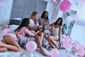 private Party. vier attraktive junge lächelnde frauen in pyjamas, die sich gegenseitig anstoßen, während sie im schlafzimmer eine schlafparty mit luftballons im ganzen raum veranstalten foto