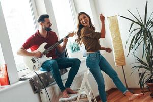 glücklicher junger Mann in Freizeitkleidung, der Gitarre spielt, während seine attraktive Freundin tanzt foto