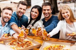 Prost auf Sie Gruppe glücklicher junger Menschen, die sich aneinander binden und Flaschen mit Bier ausstrecken, während sie im Freien stehen foto