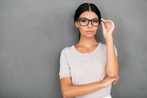 selbstbewusste Schönheit. selbstbewusste junge Geschäftsfrau, die Brillen anpasst und in die Kamera schaut, während sie vor grauem Hintergrund steht foto