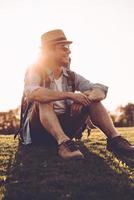 Zeit an der frischen Luft genießen. hübscher junger Mann in Fedora, der Rucksack trägt und lächelt, während er draußen auf grünem Gras sitzt foto