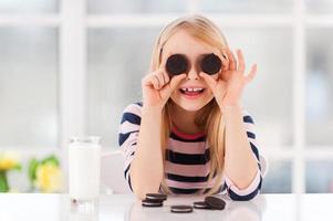 Spaß mit Keksen. Fröhliches kleines Mädchen, das die Augen mit Keksen bedeckt, während es mit einem Glas Milch darauf am Tisch sitzt foto