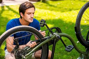 Mann repariert sein Fahrrad. Selbstbewusster junger Mann, der sein Fahrrad repariert, während er auf Gras kniet foto