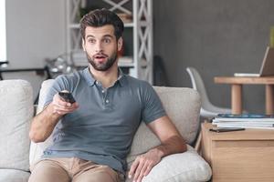 schockierende Fernsehsendung. hübscher junger mann, der fernbedienung hält und überrascht aussieht, während er zu hause auf der couch fernsieht foto