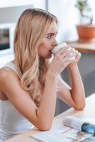 genießt ihren Morgenkaffee. Seitenansicht einer schönen jungen Frau, die Kaffee trinkt und wegschaut, während sie zu Hause in der Küche sitzt foto