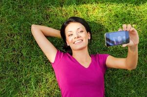 Selbstportrait machen. Blick von oben auf die schöne junge Frau, die mit ihrem Smartphone ein Selfie macht, während sie im grünen Gras liegt foto