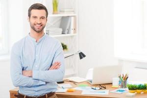 er hat einen kreativen Geist. hübscher junger Mann im Hemd, der die Arme verschränkt hält und in die Kamera lächelt, während er sich am Schreibtisch im Büro lehnt foto