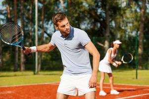 als eine Mannschaft spielen. hübscher junger Mann, der Tennisschläger hält und wegschaut, während er auf dem Tennisplatz und mit Frau im Hintergrund steht foto