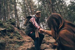 zusammen werden wir es durchstehen. Modernes junges Paar, das Händchen hält und sich beim gemeinsamen Wandern im Wald nach oben bewegt foto