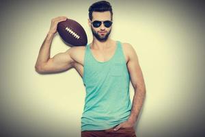 Bereit, selbstbewusster junger gutaussehender Mann mit Sonnenbrille zu spielen, der den American-Football-Ball auf der Schulter trägt und die Kamera anschaut, während er vor weißem Hintergrund steht foto