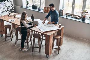 anstrengender Tag. Draufsicht auf junge Kollegen in Smart Casual, die während der Arbeit im kreativen Arbeitsbereich eine Blaupause verwenden foto