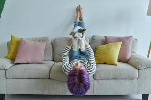 Verspielte Teenagerin, die ihren kleinen Hund trägt, während sie sich zu Hause auf der Couch entspannt foto