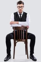 überzeugt von seinem Stil. hübscher junger Mann in Abendkleidung, der auf dem Stuhl sitzt und die Kamera anschaut, während er vor grauem Hintergrund steht foto