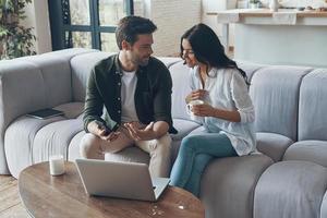 Hübscher junger Mann, der seiner Freundin ein Smartphone zeigt und lächelt, während er zu Hause auf dem Sofa sitzt foto