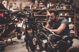 Mechaniker bei der Arbeit. Selbstbewusster junger Mann, der Motorrad in der Werkstatt repariert foto