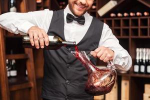 Wein in Dekanter gießen. Zugeschnittenes Bild eines selbstbewussten männlichen Sommeliers, der Rotwein in den Dekanter gießt, während er in der Nähe des Weinregals steht foto