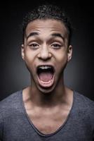 voller Emotionen. Porträt eines jungen aufgeregten afrikanischen Mannes, der mit offenem Mund in die Kamera blickt, während er vor schwarzem Hintergrund steht foto