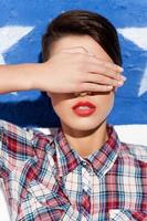 nichts sehen. schöne junge Frau mit kurzen Haaren, die vor dem Hintergrund der amerikanischen Flagge steht und die Augen mit der Hand bedeckt foto