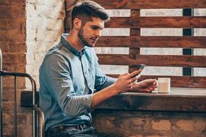 ein paar minuten für kaffee und kurze nachricht. Seitenansicht eines hübschen, nachdenklichen jungen Mannes, der ein Smartphone hält und es betrachtet, während er mit einer Kaffeetasse in der Hand in der Nähe des Fensters im Loft-Interieur sitzt foto