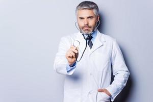 medizinische Untersuchung. selbstbewusster reifer Arzt mit grauem Haar, der Sie mit Stethoskop untersucht, während er vor grauem Hintergrund steht foto