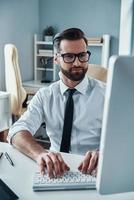 moderner junger Mann in Abendkleidung, der am Computer arbeitet, während er im Büro sitzt foto