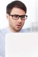 schockierende Nachrichten. überraschter junger mann in hemd und krawatte, der auf den laptop schaut, während er an seinem arbeitsplatz sitzt foto
