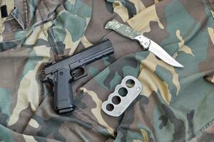 Pistole liegt mit Schlagring und Messer auf Tarnuniform aus nächster Nähe. Konzept der Plünderung und des Waffenhandels foto